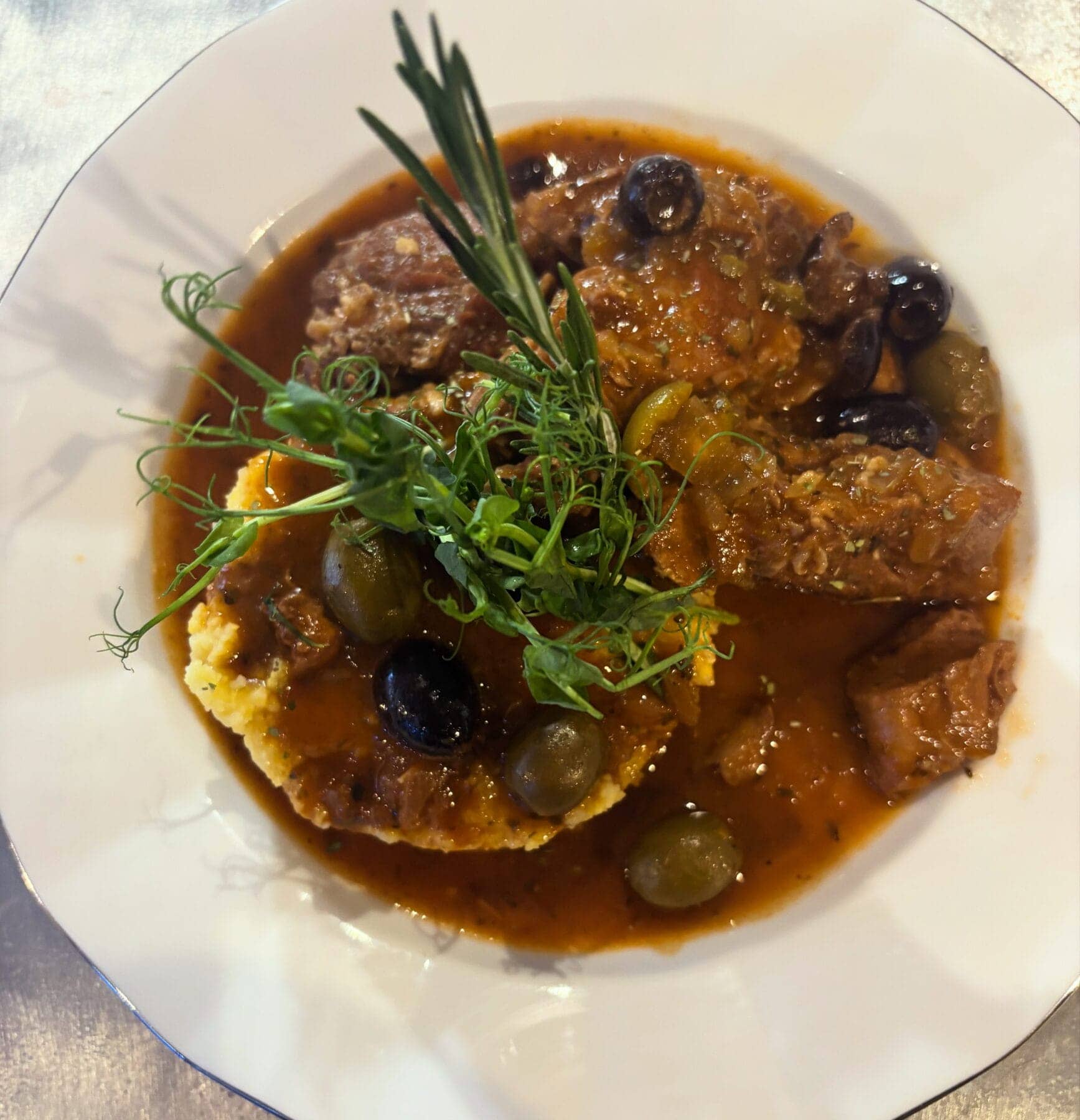 Plat mijoté de veau en sauce aux olives vertes et noires, servi sur une purée dans une assiette bleue, un plat maison du Bistrot Napoléon à emporter.